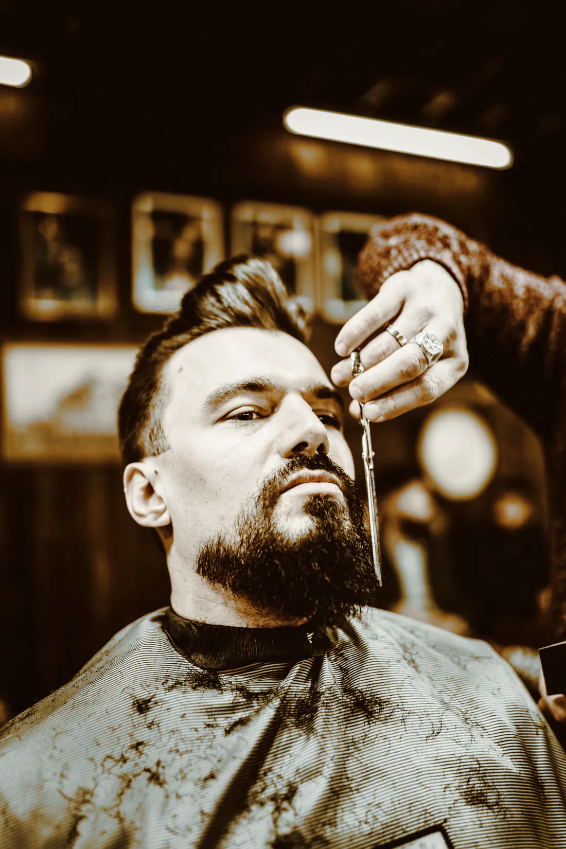 A barber trimming a client’s dark beard with scissors, framed pictures on the wall behind