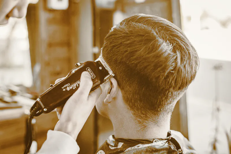 A barber tapering the back of a client’s haircut with clippers beside a bright window