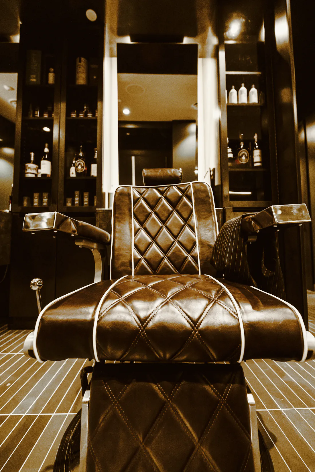 A brown leather barber chair with diamond stitching in front of a lit mirror