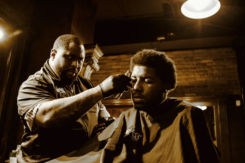 A barber in black gloves lining up a young man’s hair under warm evening light