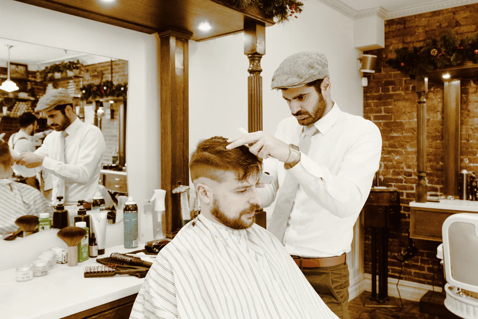 A barber in a flat cap and white shirt checking a seated client’s haircut in a bright timber shop