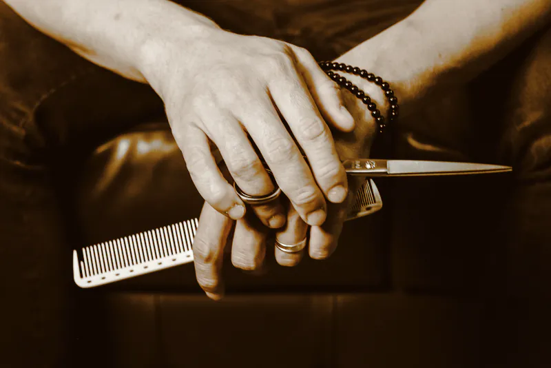 A barber’s hands resting on a leather chair, holding a comb and scissors