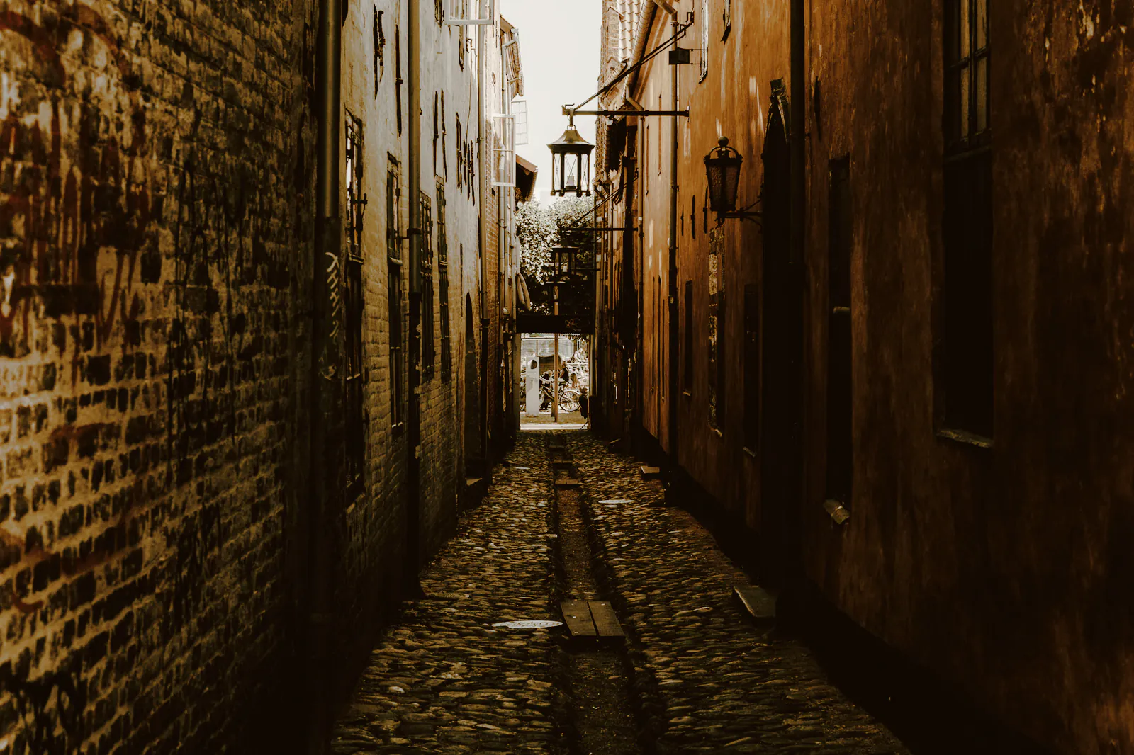 A narrow cobbled lane between old brick walls, a lantern hanging over it