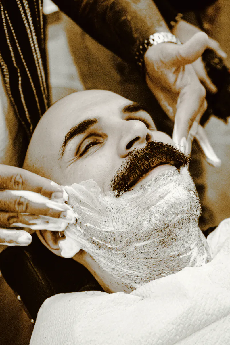 A smiling man with a moustache having his cheeks lathered for a shave