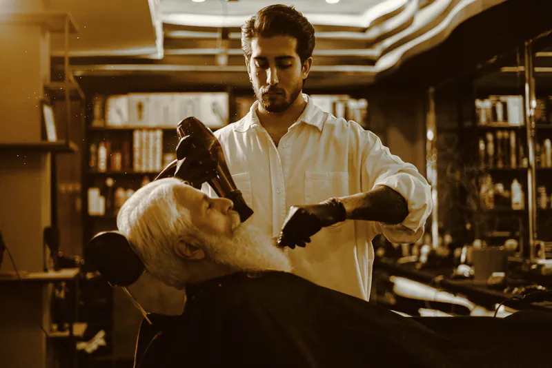 A barber in a white shirt and black gloves tending an older client’s white beard in a dark shop lined with books
