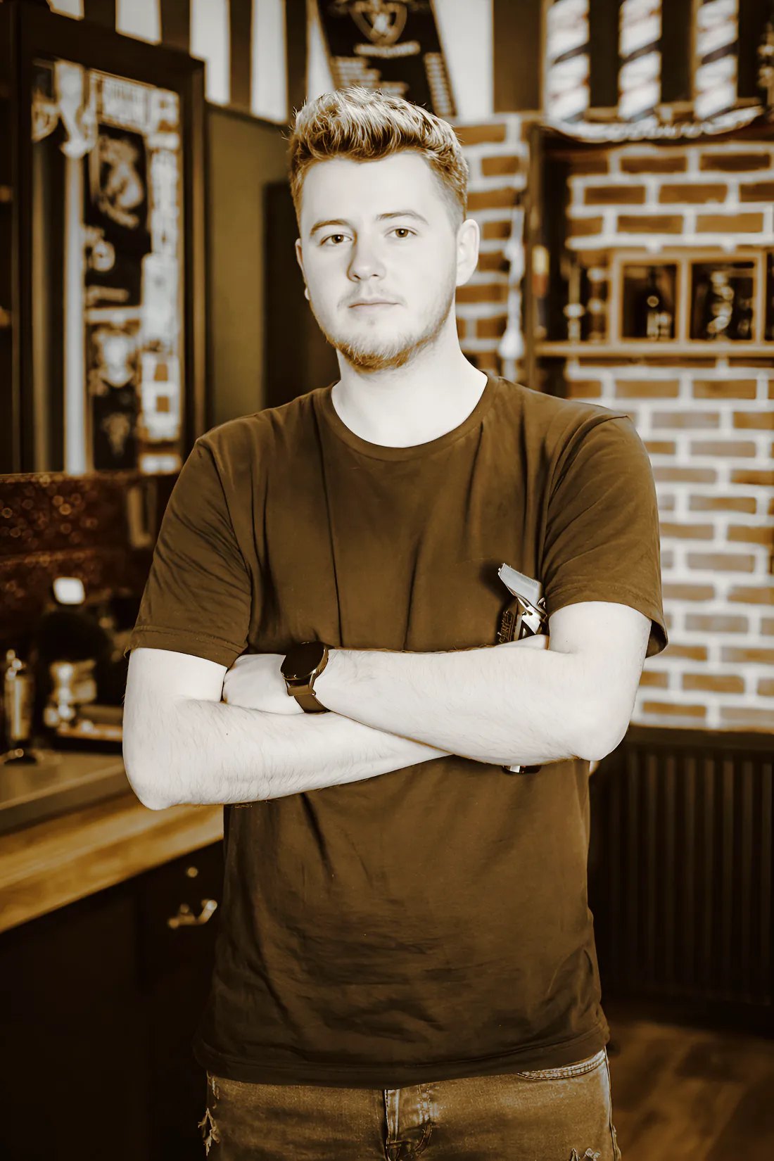 A young barber with his arms crossed in front of a brick wall and striped poles