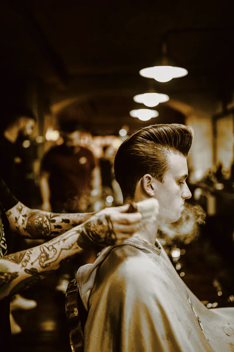 Tattooed arms brushing cut hair from the neck of a client with a tall pompadour