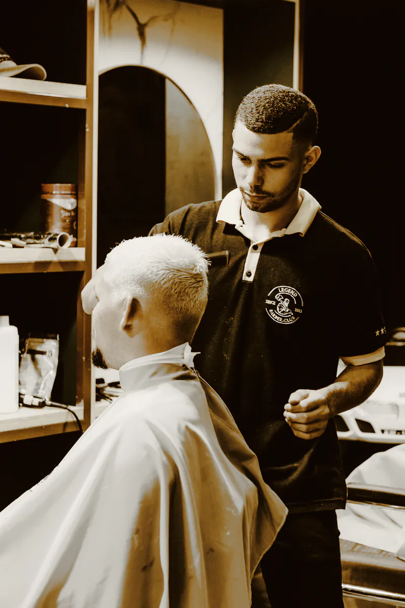 A barber in a black polo shirt shaping a client’s bleached crop
