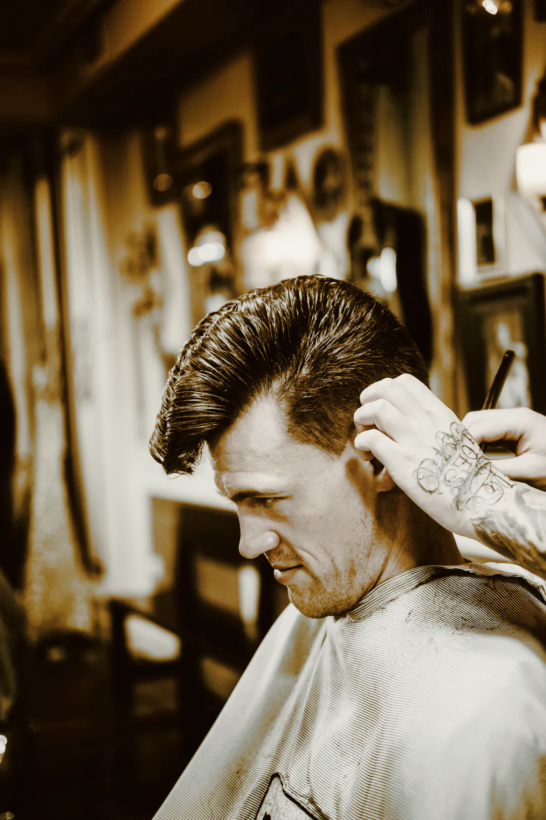 A client with a dark pompadour looking down while a tattooed hand works at his temple