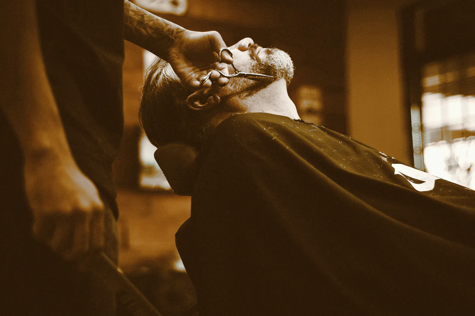 A barber trimming a reclining client’s grey beard with scissors in low light