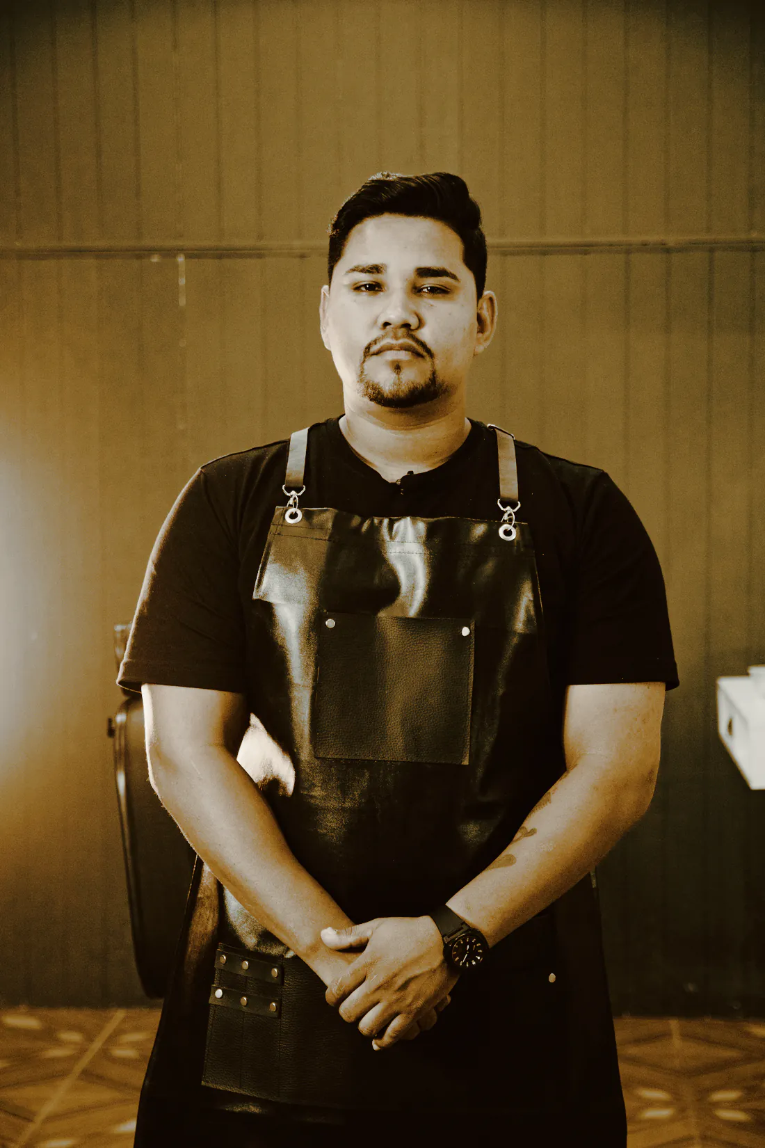 A barber in a black leather apron standing with his hands clasped