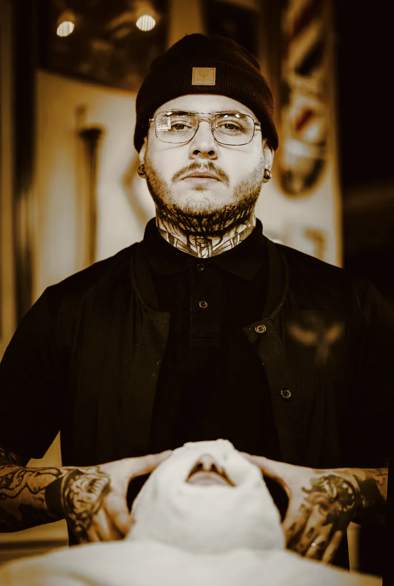 A tattooed barber in a beanie and glasses, his hands resting on a hot towel over a client’s face