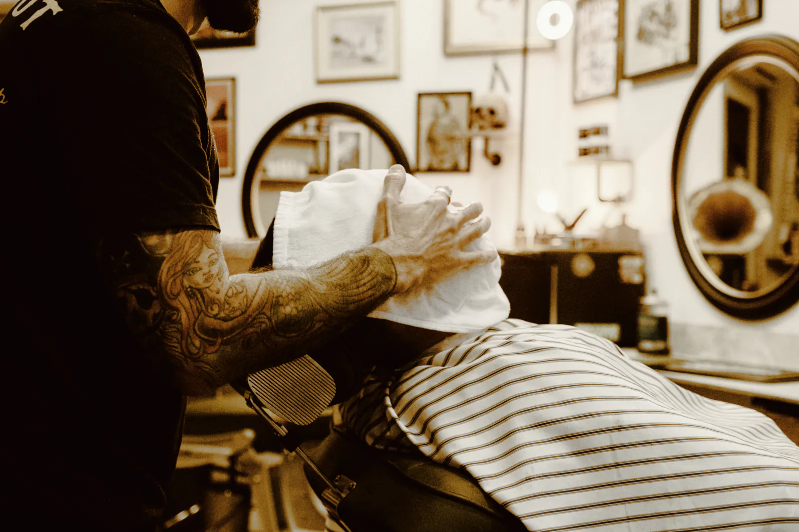 A tattooed barber pressing a hot towel to a reclining client’s face in a shop hung with framed pictures