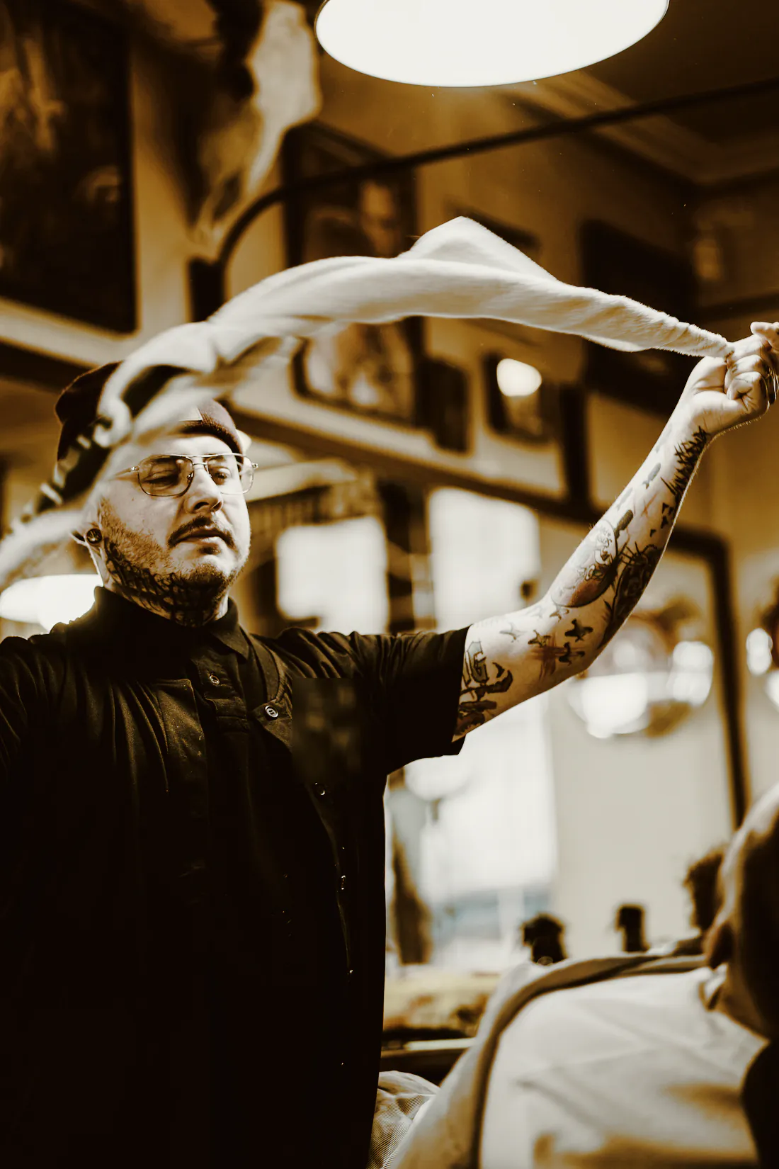 A tattooed barber in glasses shaking out a hot towel in a shop hung with framed pictures